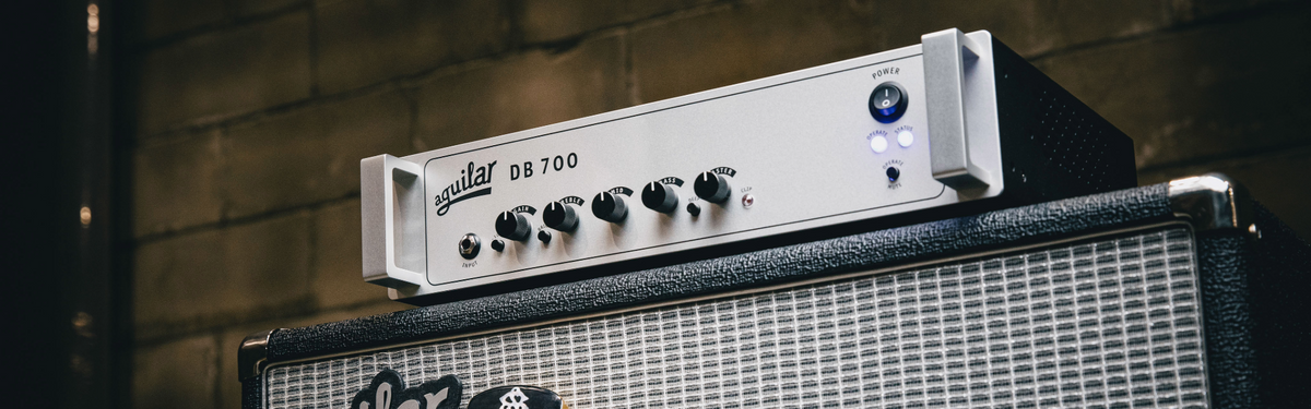 A silver and black guitar amplifier head labeled 'Guitar DB 700' resting on top of a black speaker cabinet against a dark brick wall.